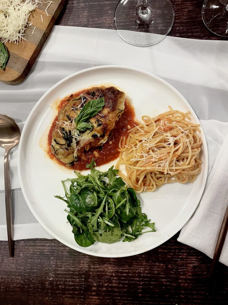 easy weeknight chicken parmesan with spagthetti and arugula salad on a white plate with a rose gold spoon and the stem of two wine glasses.