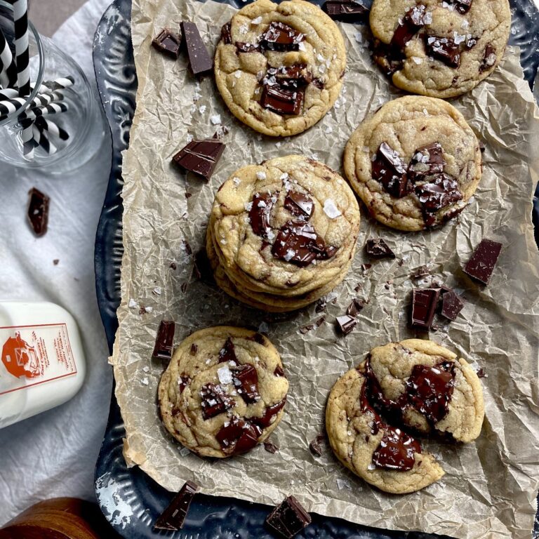 brown butter sea salt chocolate chip cookies on parchment paper with chopped chocolate and flaked sea salt sprinkled all around. Glass bottle of sherman's farm milk.