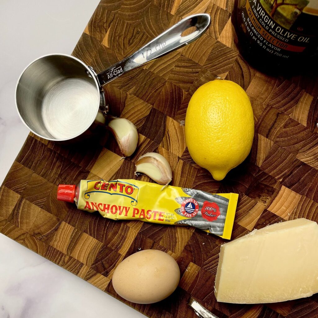 A whole lemon, 2 cloves of garlic, tube of anchovy paste, an egg, block of parmesan, bottle of olive oil and a stainless steal 1/2 cup measuring cup on a wooden butchers block.