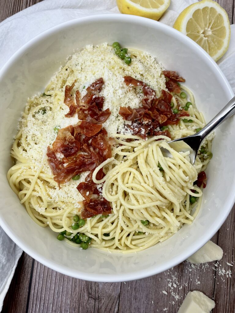prosciutto lemon pasta in a white serving bowl with a forkful of spaghetti twisted onto a fork. Topped with fresh grated romano and crispy prosciutto.