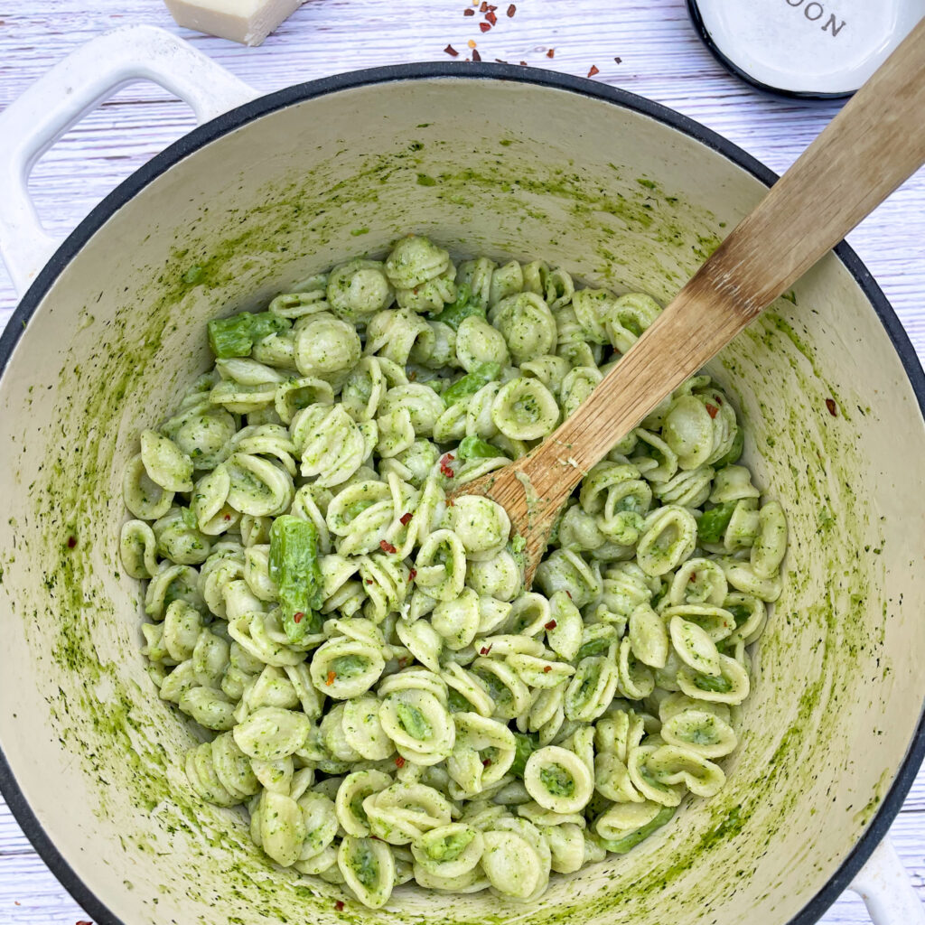 orecchiette con broccoli in a white enameled cast iron pot with a bamboo spoon in the middle. Placed on a white picnic table with sprinkled chili flakes around it.
