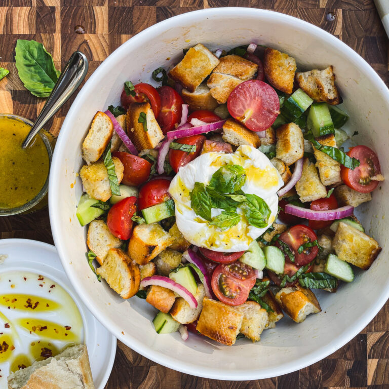 Panzanella toscana with grilled bread and burrata in a white serving bowl with basil leaves on top of the burrata. Dish is on top of a butcher block with a small glass of vinaigrette and a bread dipping dish with extra virgin olive oil and red pepper flakes.