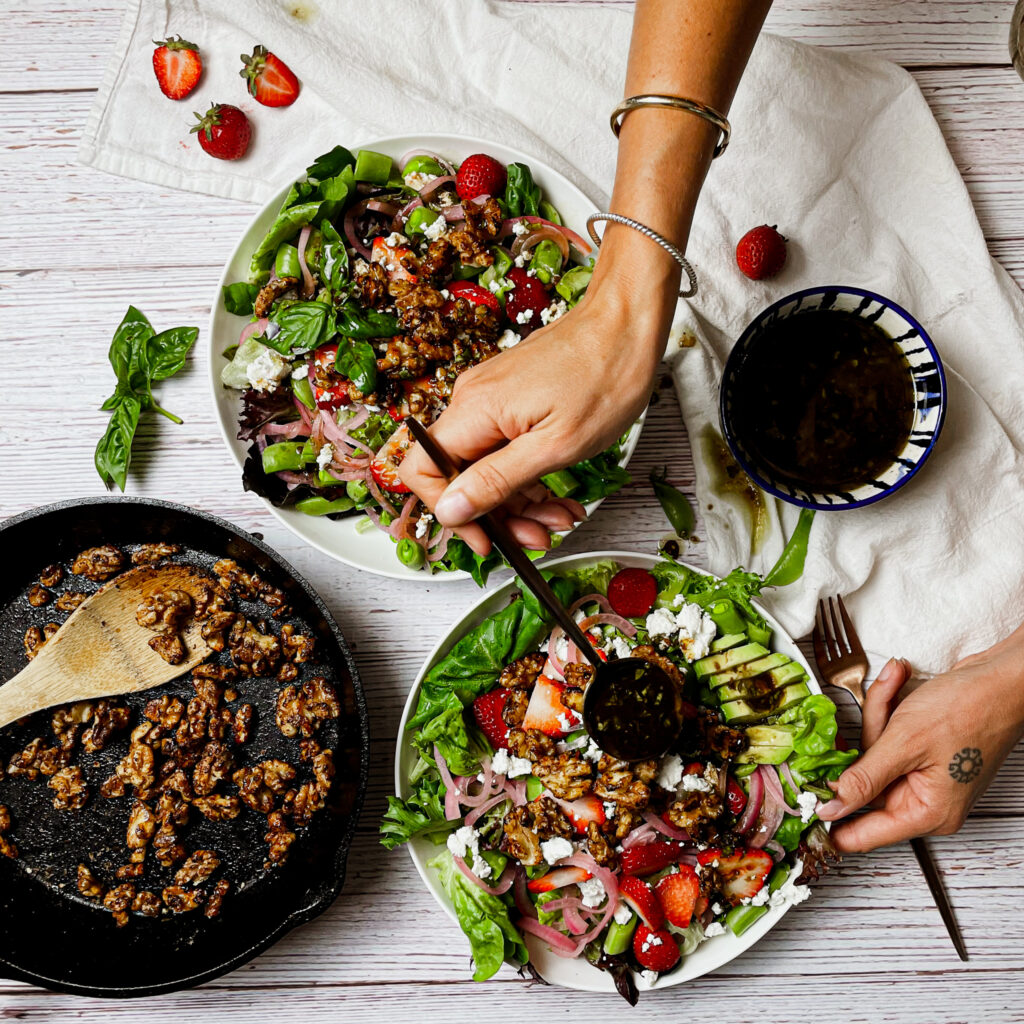a cast iron pan on a white picnic table with maple toasted walnuts, a hand spooning basil vinaigrette onto a bowl of the strawberry walnut goat cheese salad.