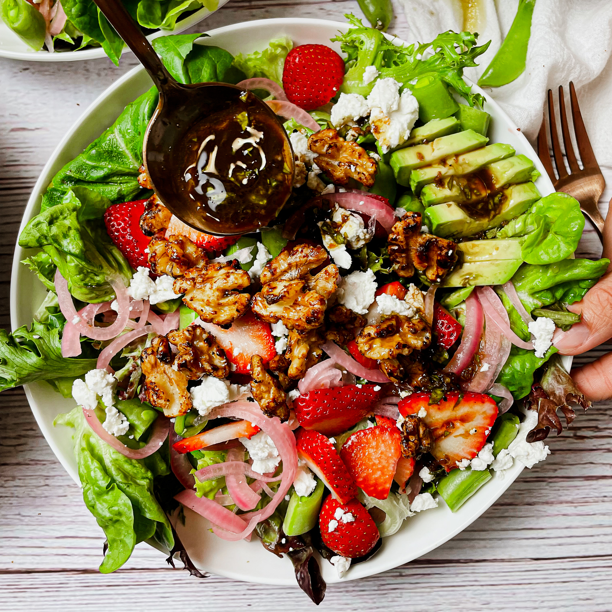 close up image of rose gold spoon ladling the dressing onto a bowl of the strawberry walnut goat cheese salad in a white salad bowl