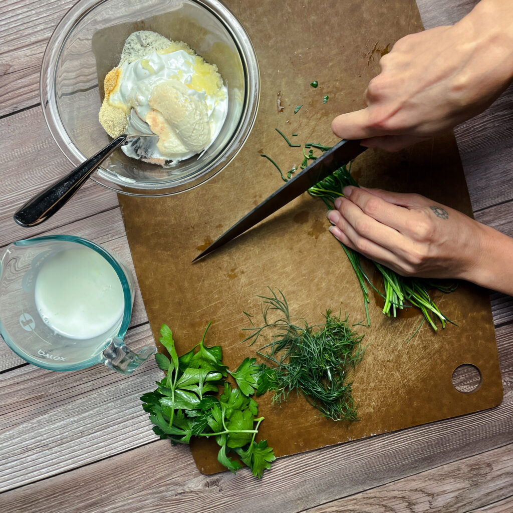 cutting board with fresh herbs, hand using a chefs knife chopping the chives to put in the glass bowl that has ranch dressing ingredients; sour cream, mayo, milk, ranch seasoning.
