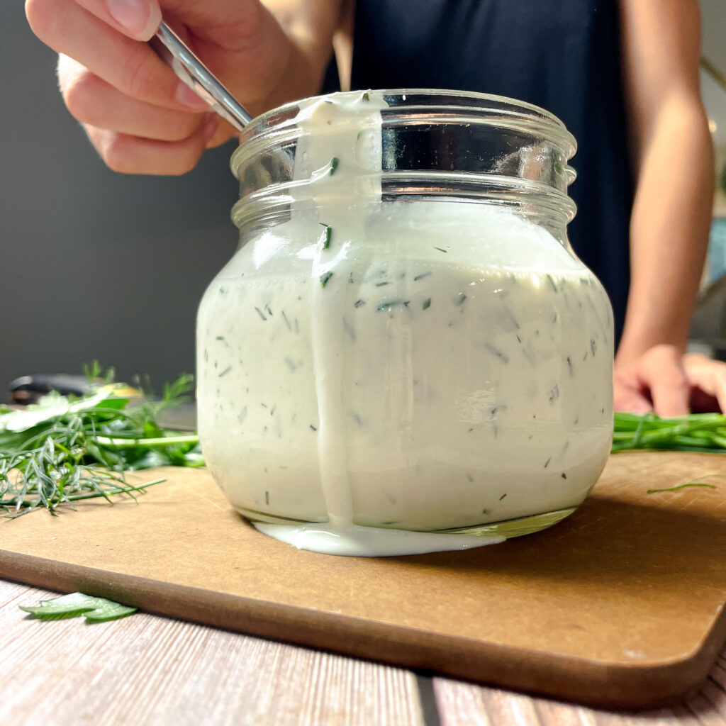 Close up image of ason jar of fresh homemade ranch dressing, with a pile of fresh herbs next to it.