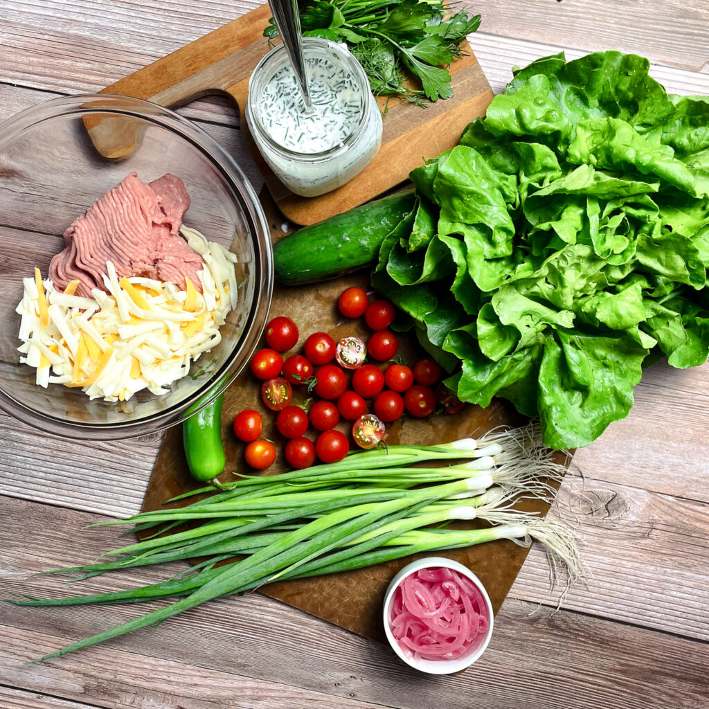 Fresh farm vegetable ingredients for this recipe on a cutting board, bibb lettuce, grape tomatoes, green onions, cucumber, a bunch of fresh herbs, mason jar of ranch dressing with a spoon in it, a small ramekin of pickled onions, a glass bowl with ground turkey, shredded cheese, and ranch seasoning, not yet mixed together. On top of a picnic table.