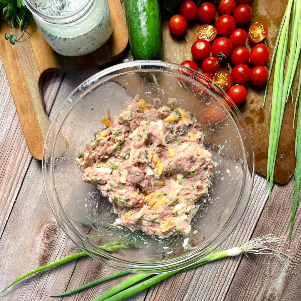 jalapeño ranch turkey burger mixture in a glass bowl, ready to be prepared into patties, on a picnic table, with green onions laying next to it. Glass bowl with turkey mixture is sitting next to a cutting board with grape tomatoes, a cucumber, and mason jar full of homemade ranch dressing.