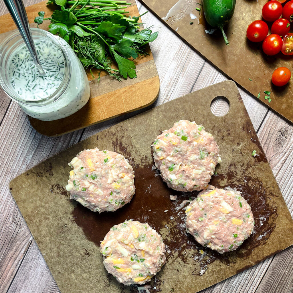 Four jalapeño ranch turkey burger patties on cutting board, prepared to be grilled. Cutting board is on a picnic table beside a mason jar of fresh homemade ranch dressing, with a pile of fresh herbs next to it.