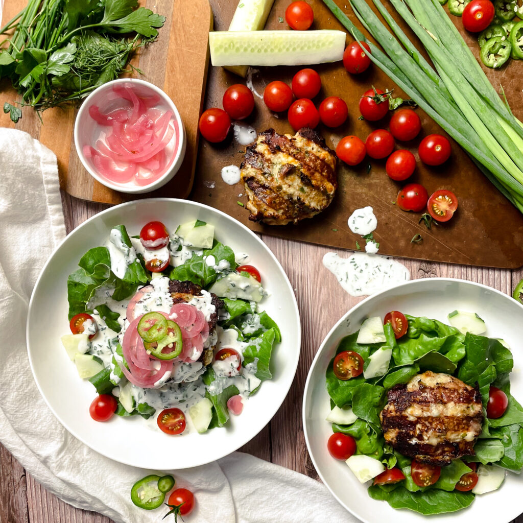 two jalapeño ranch turkey burger on top of a salads in white salad bowls. One is topped with ranch dressing, then pickled onions and fresh jalapeños. A cutting board covered in garden vegetables is sitting beside the salads.
