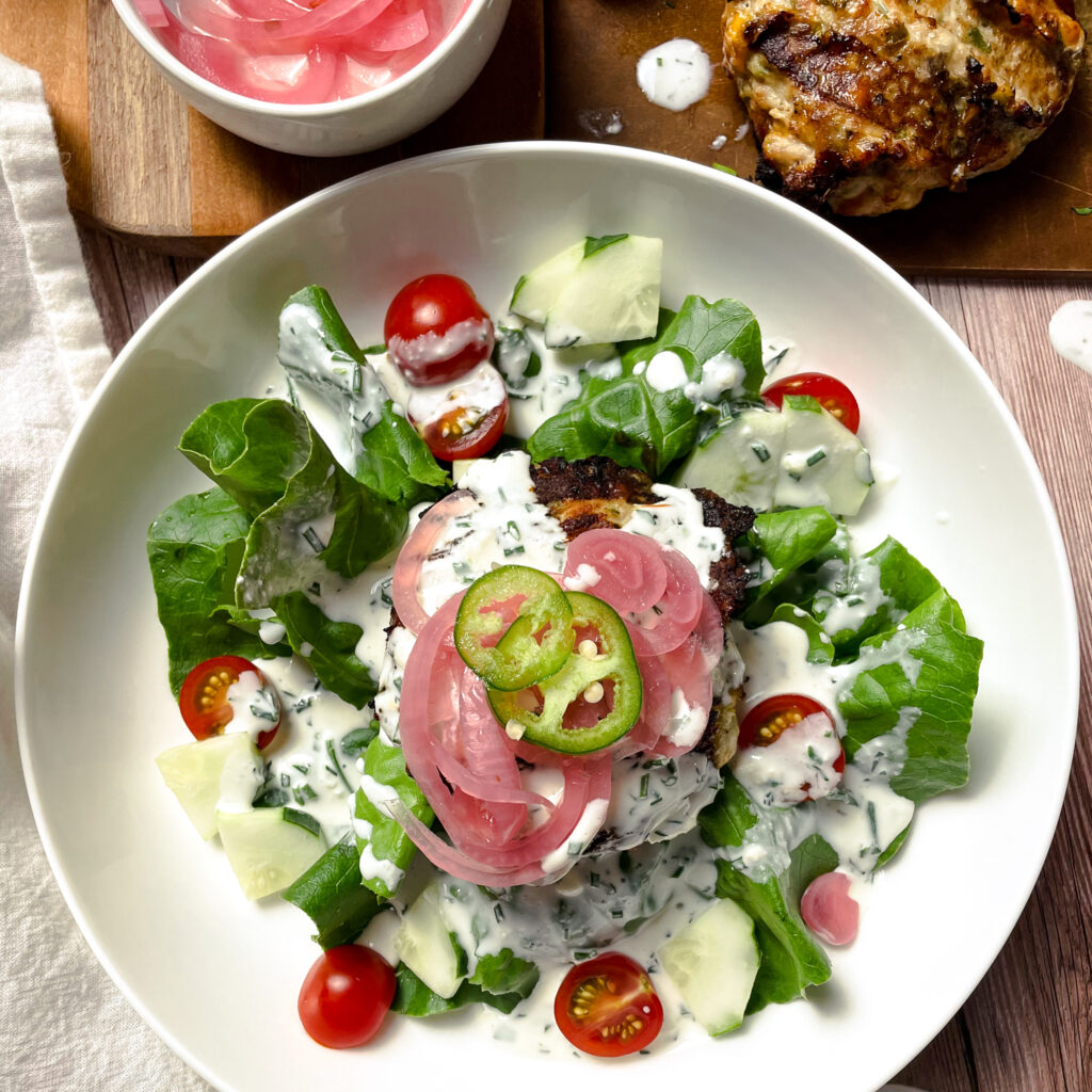 a close up image of a jalapeño ranch turkey burger sitting on a garden salad, in a while salad bowl. The burger is topped with homemade ranch dressing, purple pickled onions, and fresh sliced jalapeños.