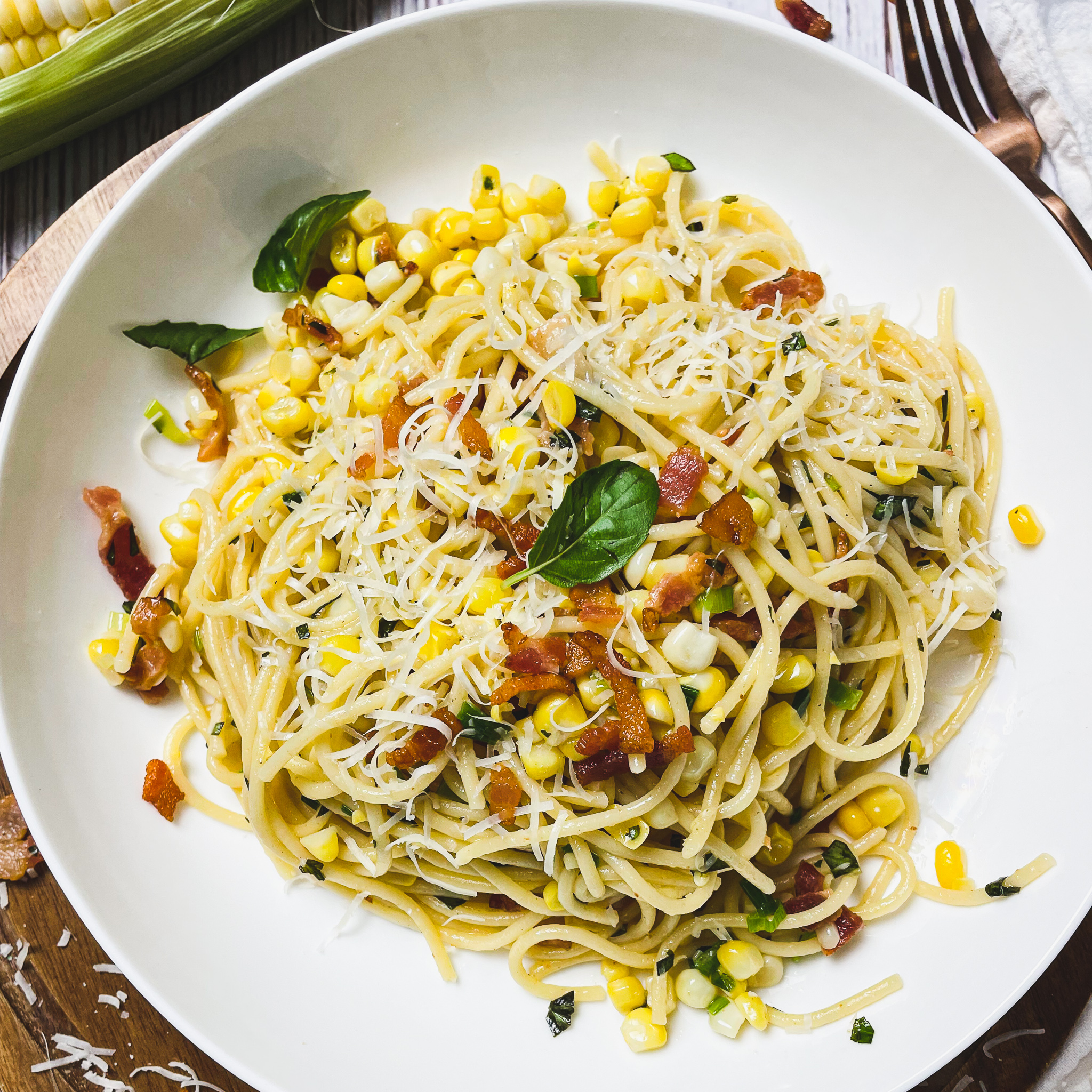 bacon and sweet corn pasta in a white pasta bowl with a basil leaf on top. Half peeled corn on the cob sitting on the table.