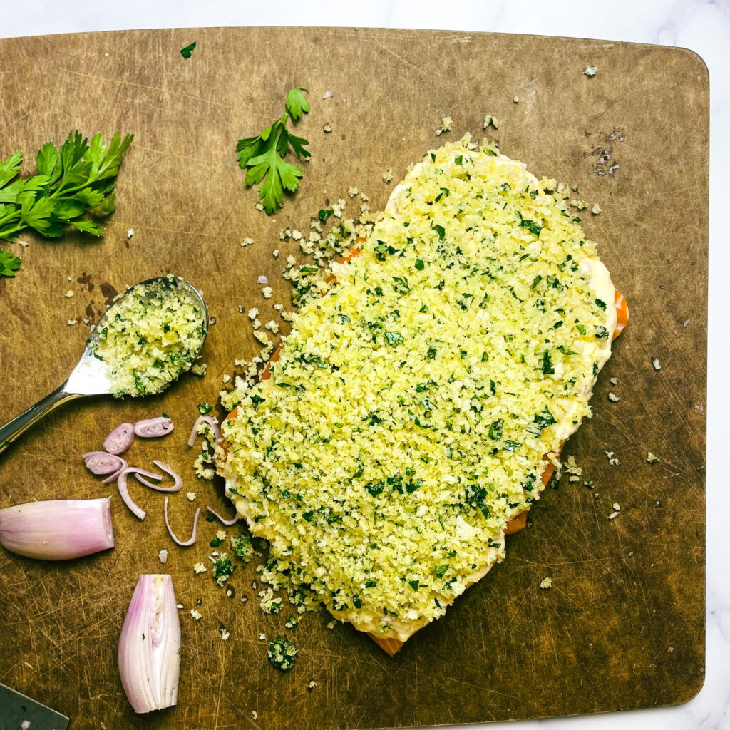 a salmon filet covered in the maple dijon cream sauce with panko crust on top, sitting on a cutting board with a sliced shallot and parsley