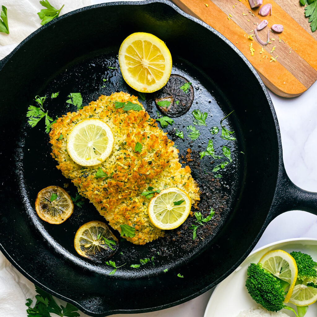 cooked maple dijon panko crusted salmon in a cast iron skillet on a kitchen table for serving, with sliced lemon and fresh parsley in the pan and a white plate of fresh steamed broccoli