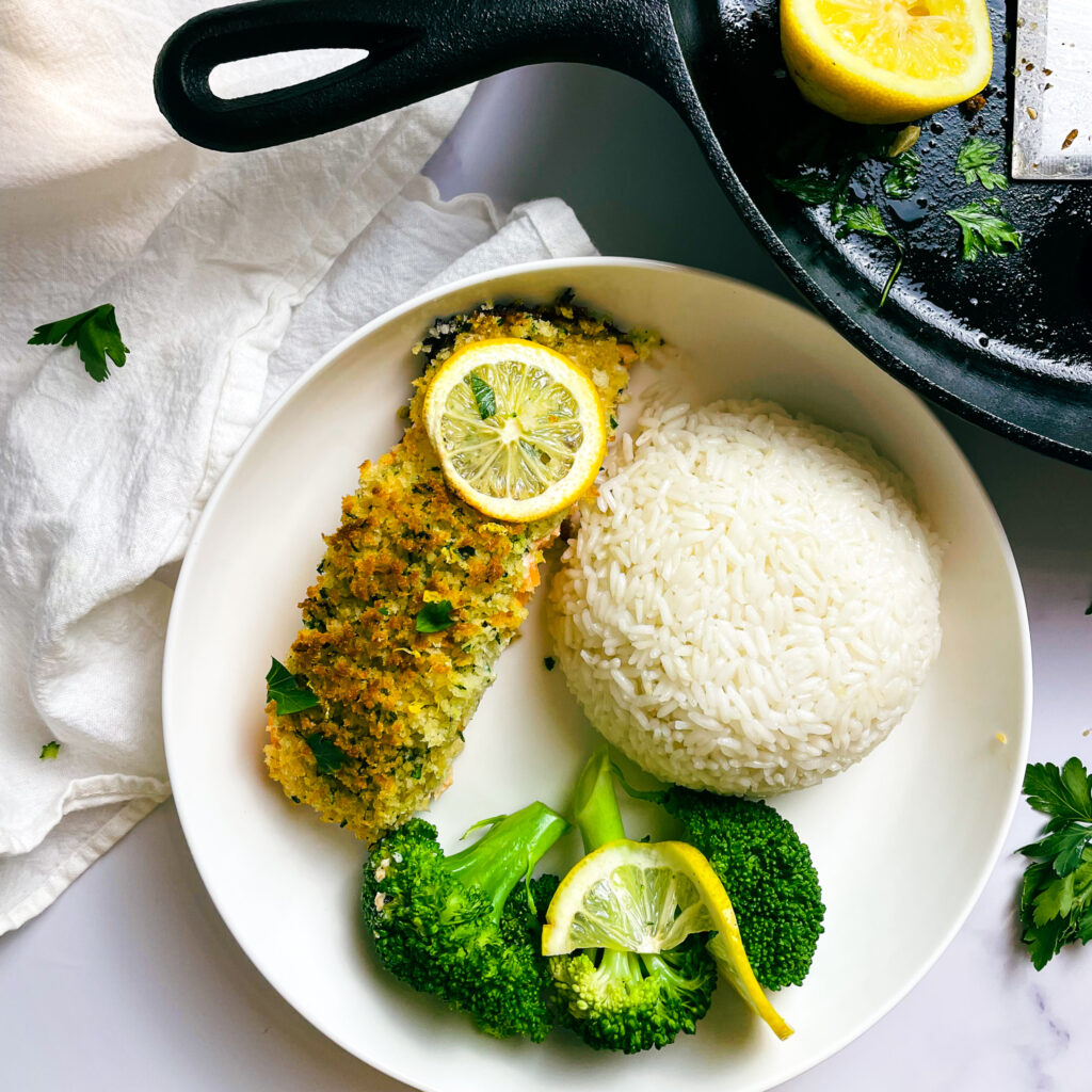 A sliced of the maple dijon panko crusted salmon on a plate with a scoop of white rice and streamed broccoli with a slice of fresh lemon on top.