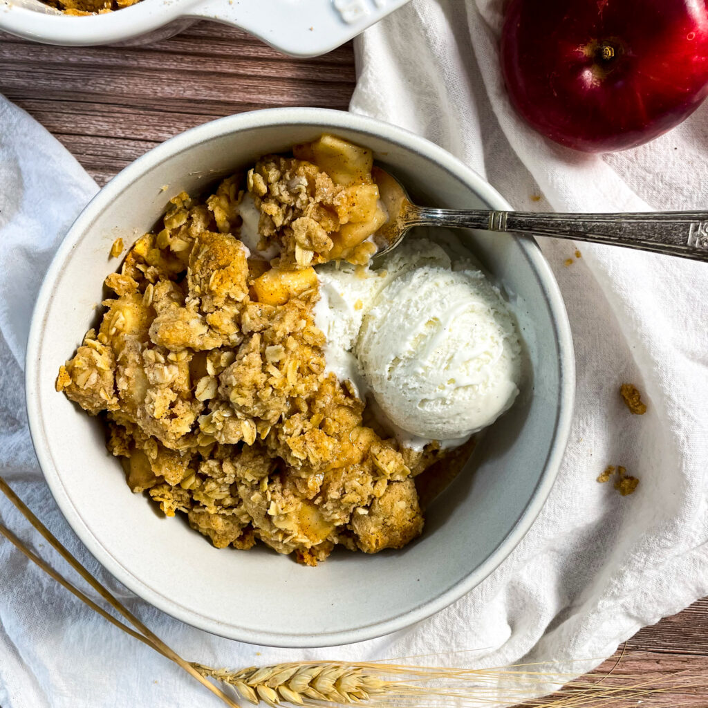 a serving of apple crisp with McIntosh apples with a scoop on top in a white bowl with a small antique spoon