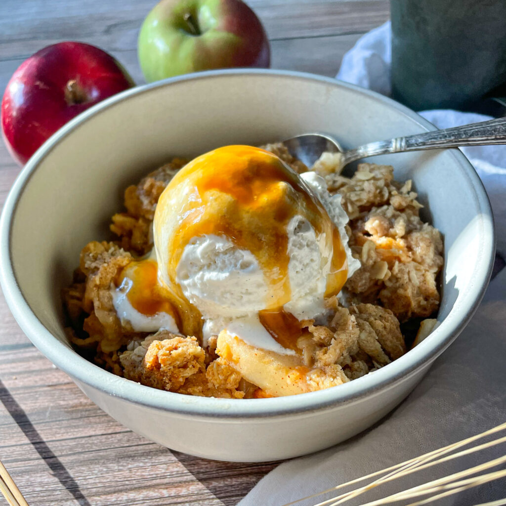 a serving of apple crisp with McIntosh apples with a scoop on top in a white bowl with a small antique spoon with caramel sauce poured on top of the ice cream
