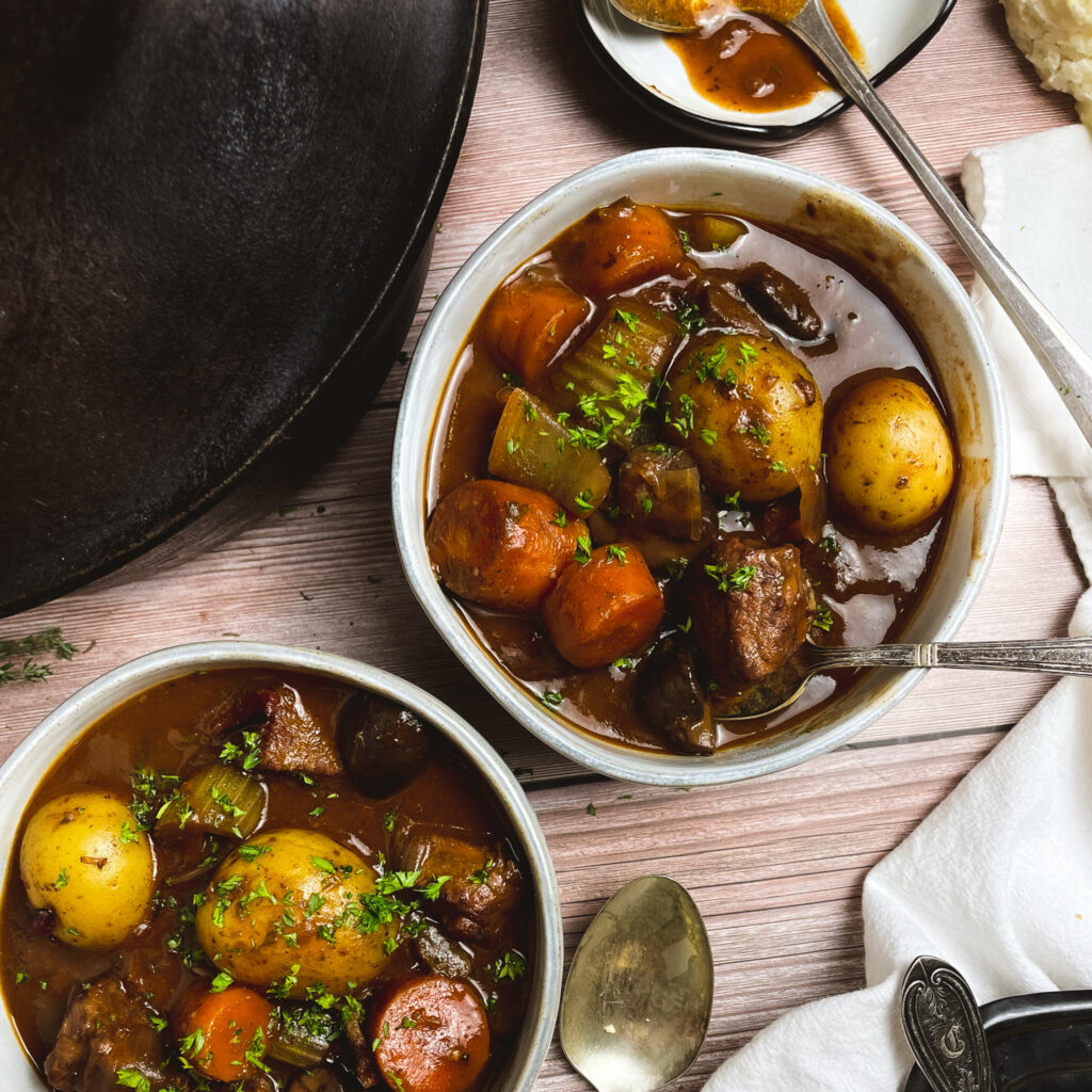 Two bowls of stovetop beef stew next to a cast iron dutch oven pot.
