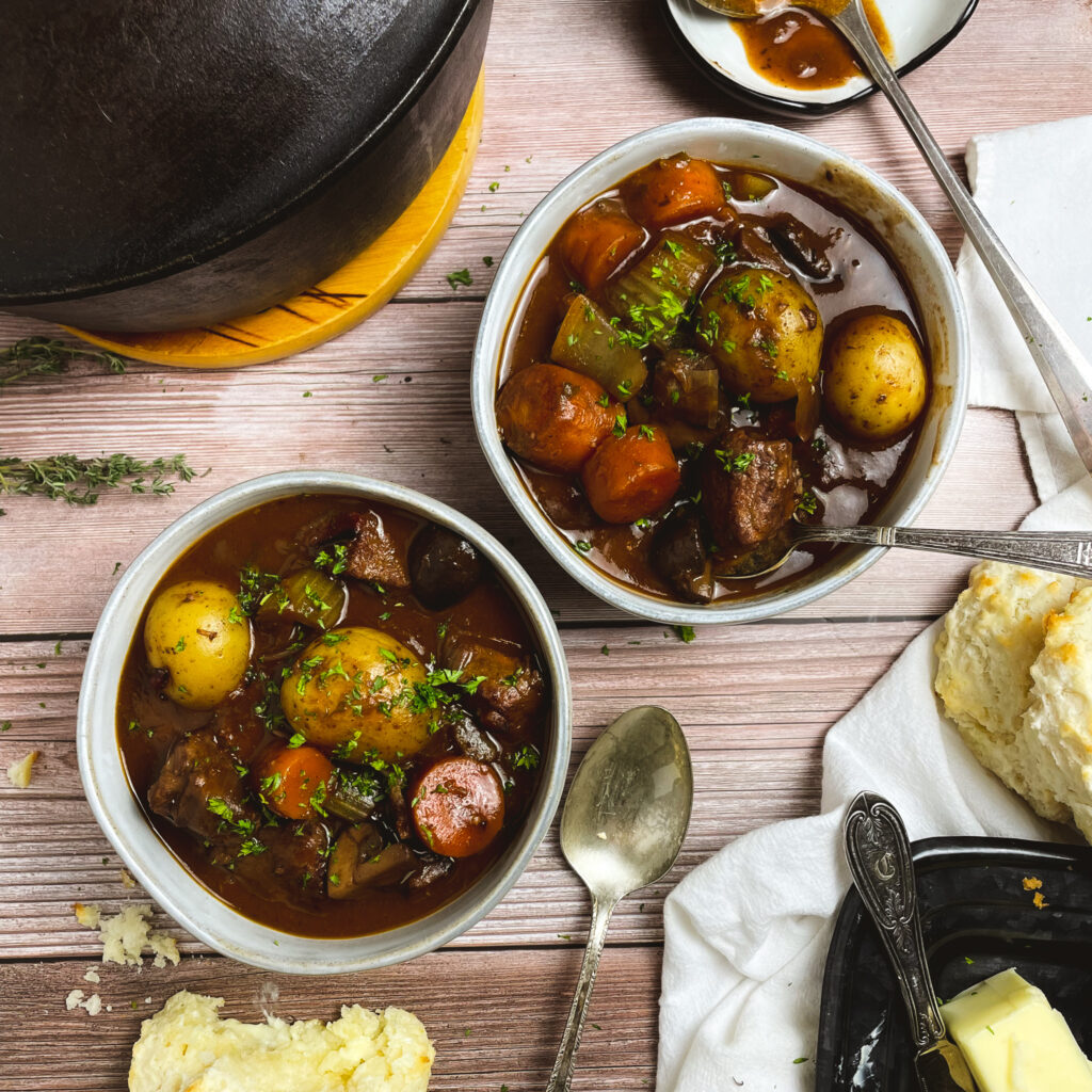 Two bowls of stovetop beef stew next to a cast iron dutch oven pot.