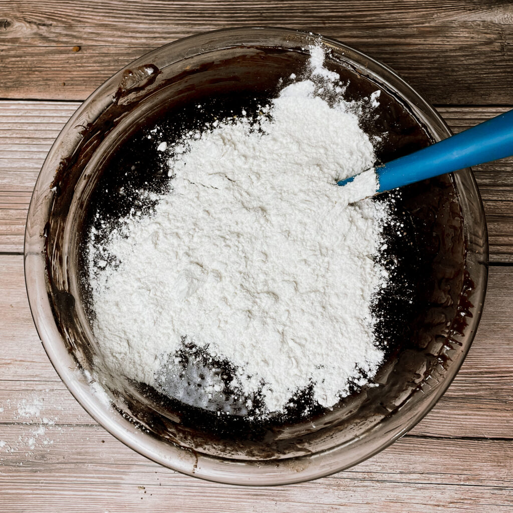 brownie batter in a glass mixing bowl with the flour sifted on top waiting to be folded in with rubber spatula.