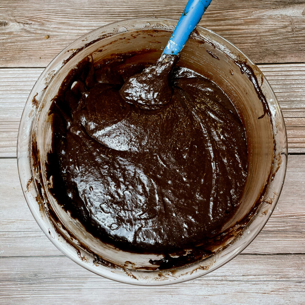 All brownie batter ingredients mixed together in a glass mixing bowl, with a blue rubber spatula.