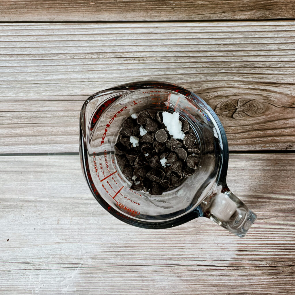 Semi sweet chocolate chips in a glass pyrex measuring cup with a little scoop of coconut oil on top.