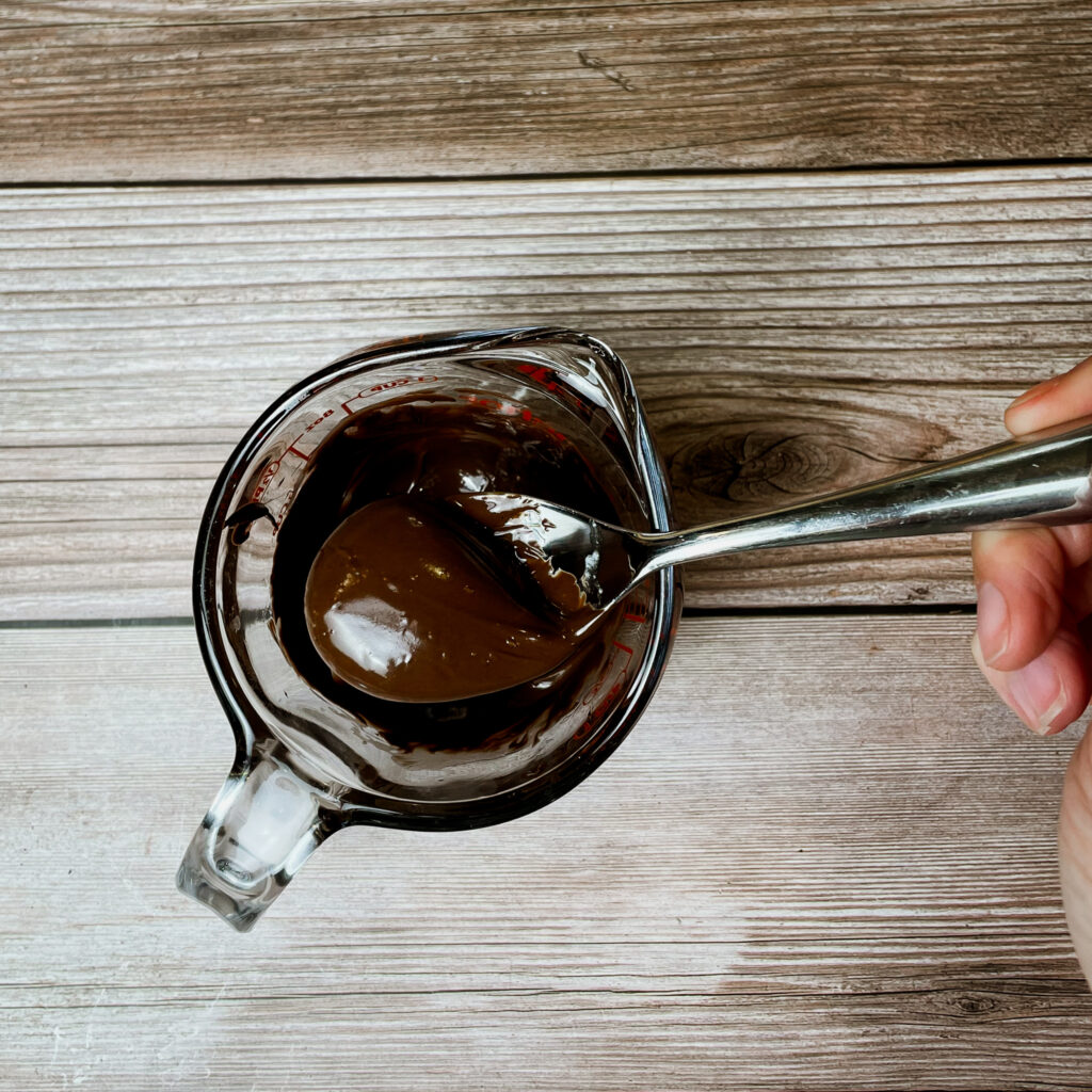 Melted semisweet chocolate in a glass pyrex measuring cup with a metal spoon, on a wooden countertop.