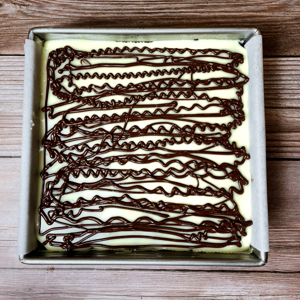 brownies in an 8x8 metal baking pan, topped with the melted chocolate, drizzled with melted semisweet chocolate on top.