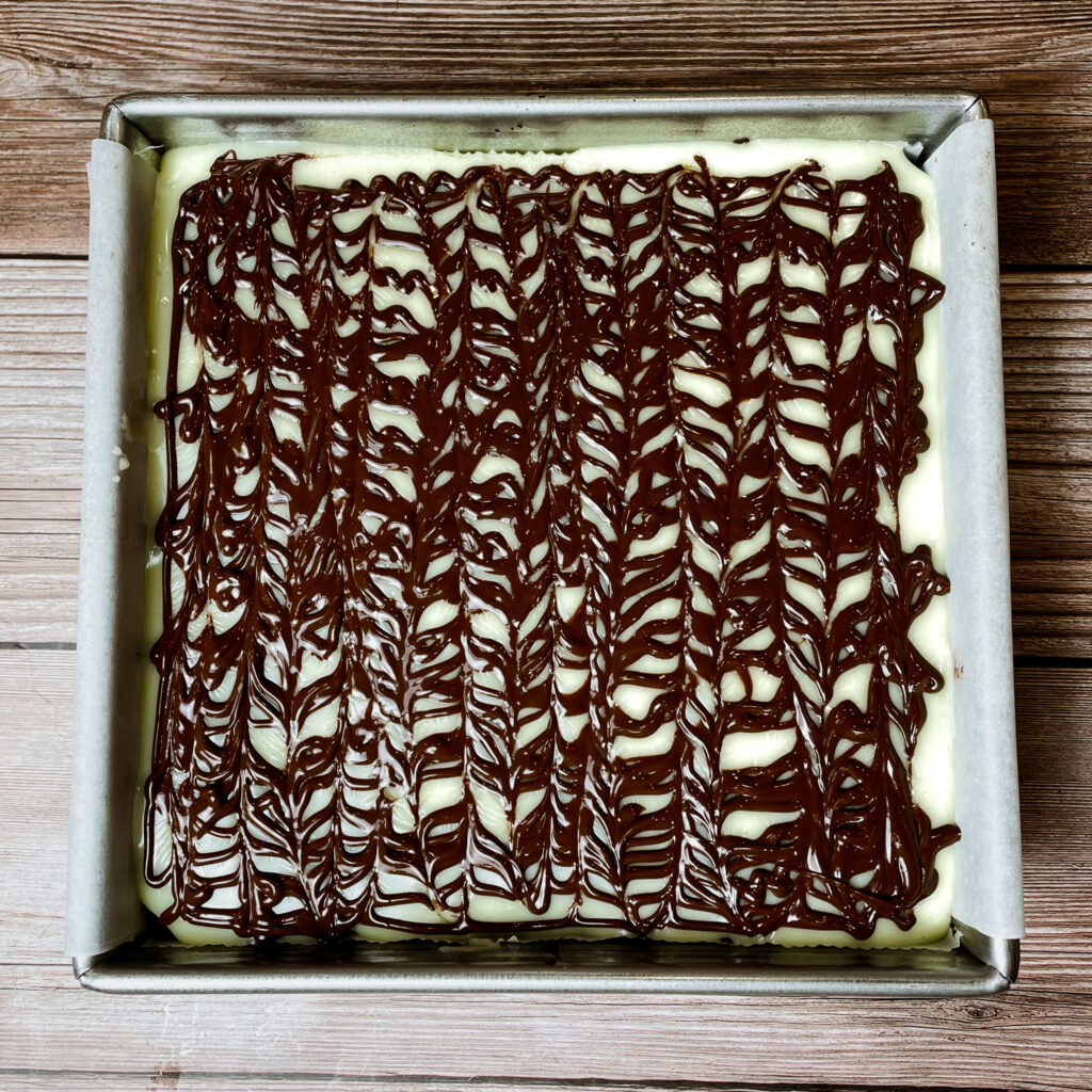 brownies in an 8x8 metal baking pan, topped with the melted chocolate, drizzled with melted semisweet chocolate on top, made into swirls.