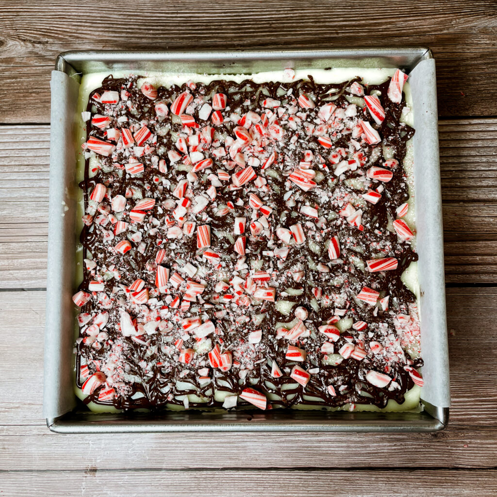 Peppermint bark brownies in an 8x8 metal baking pan topped with melted white chocolate, semi sweet chocolate and sprinkled with candy cane pieces.