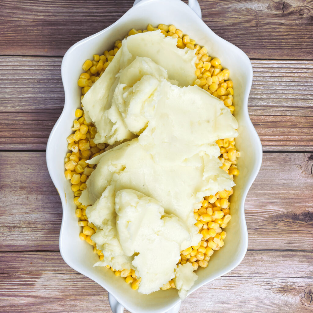 Mashed potatoes dolloped onto corn layer in white casserole dish