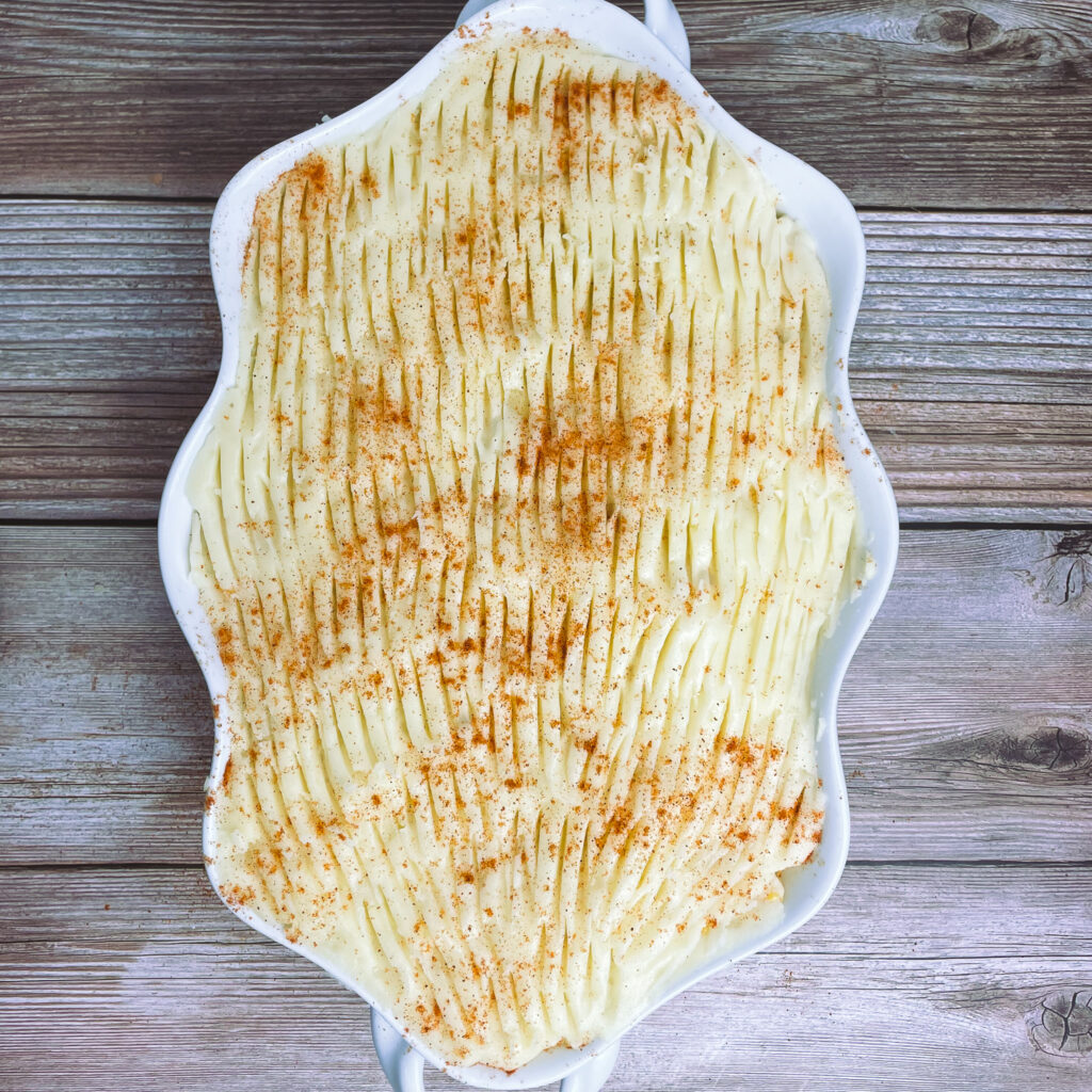 Mashed potato layer with fork marks on the top for crisping, sprinkled with paprika, in a white casserole dish