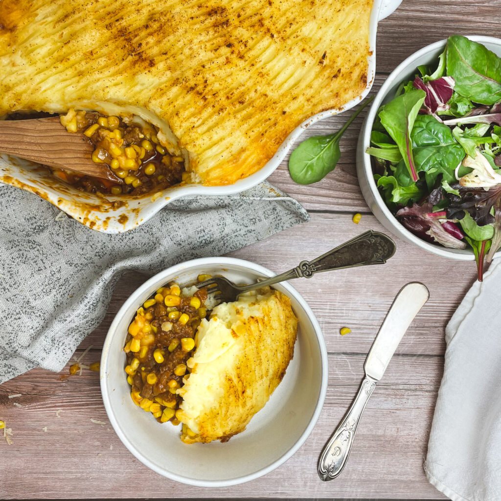 a serving of shepherd's pie with corn in a small white bowl next to a side salad and a white casserole dish with the shepherd's pie and a serving spoon.