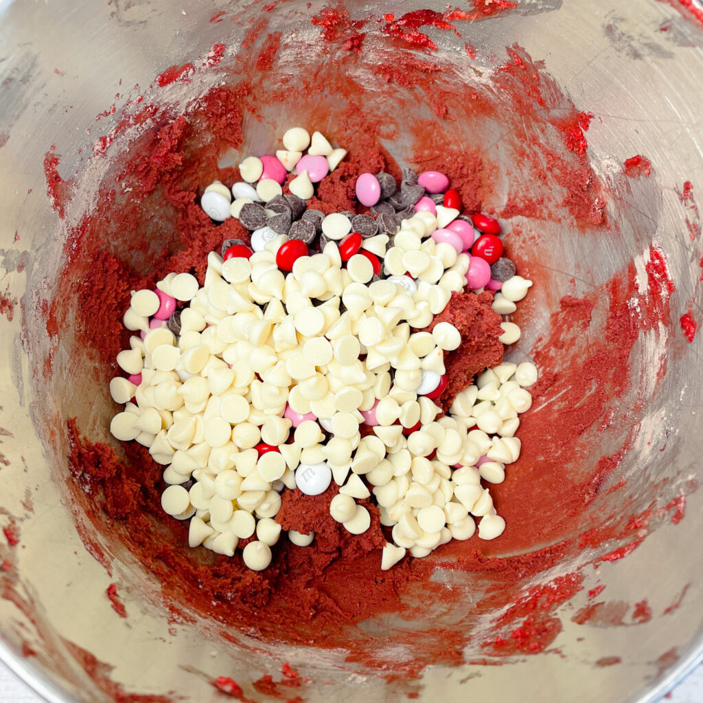 Red velvet crumbl cookie dough mixed together in a mixing bowl with M&Ms, and white chocolate chips poured on top.
