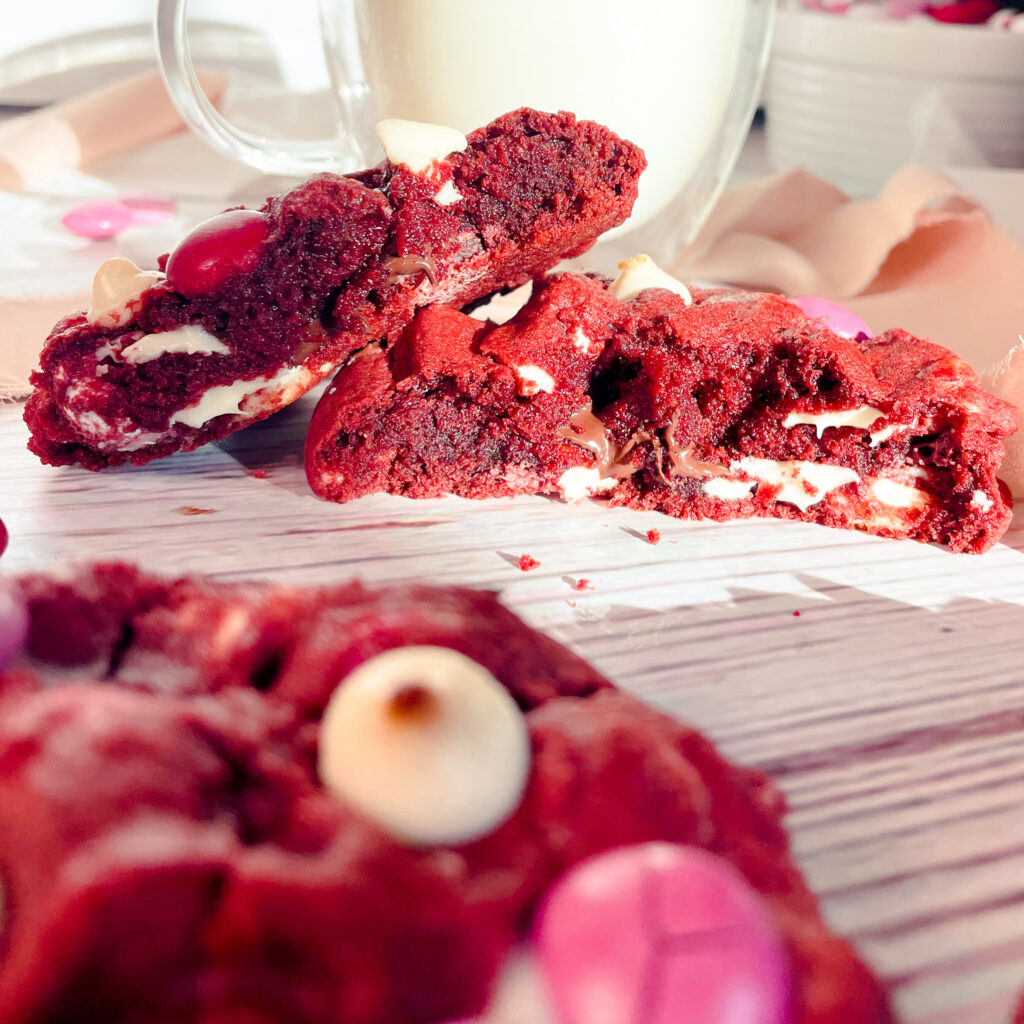 a closeup view of the a red velvet crumbl cookie split in half in front of a glass of milk.
