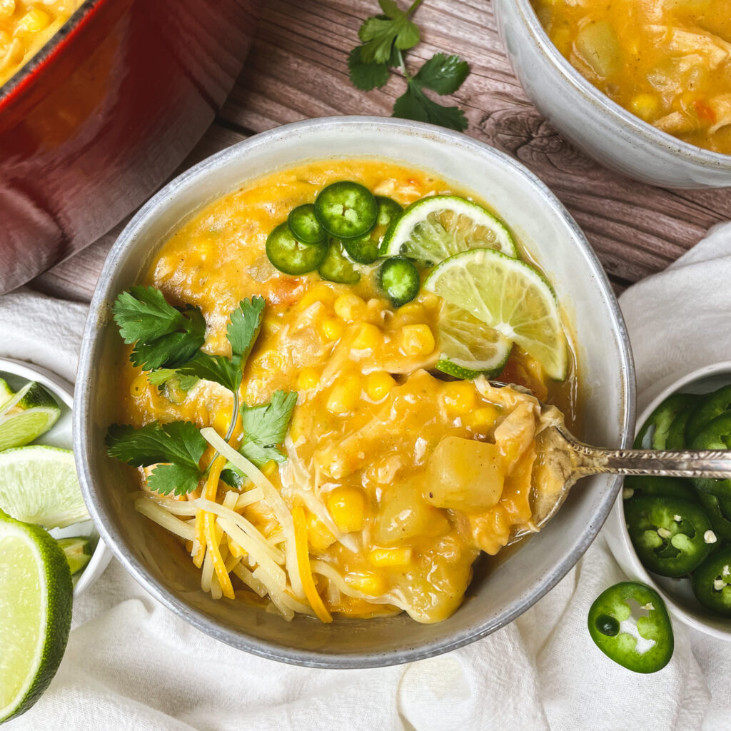 a serving of cajun chicken corn chowder in a white bowl toppedd with grated cheddar, sliced limes, sliced jalapeños, and chopped cilantro, with a spoon, on a white linen cloth.