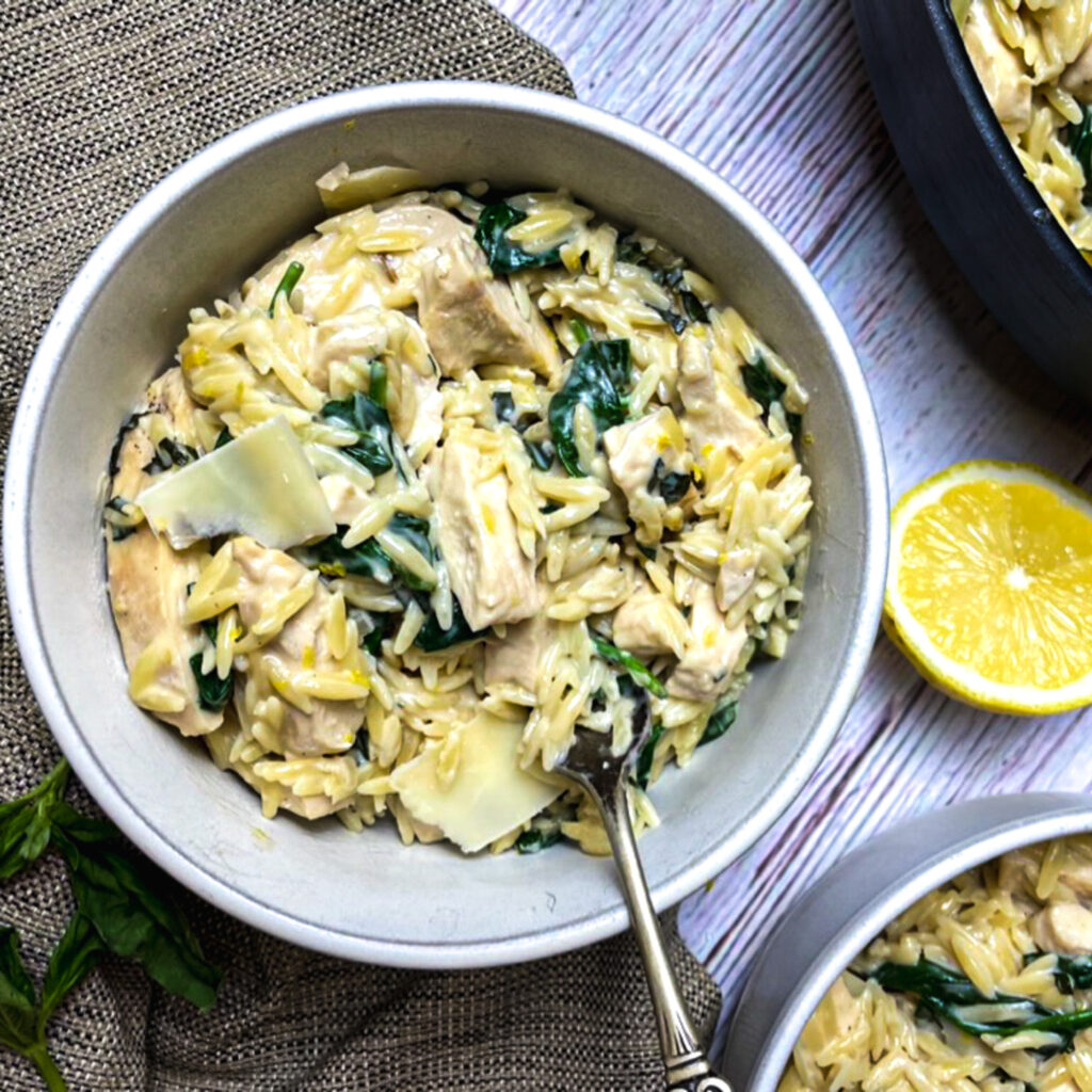 A serving of one pan lemon chicken orzo in a white bowl with shaved parmesan on top and fork.