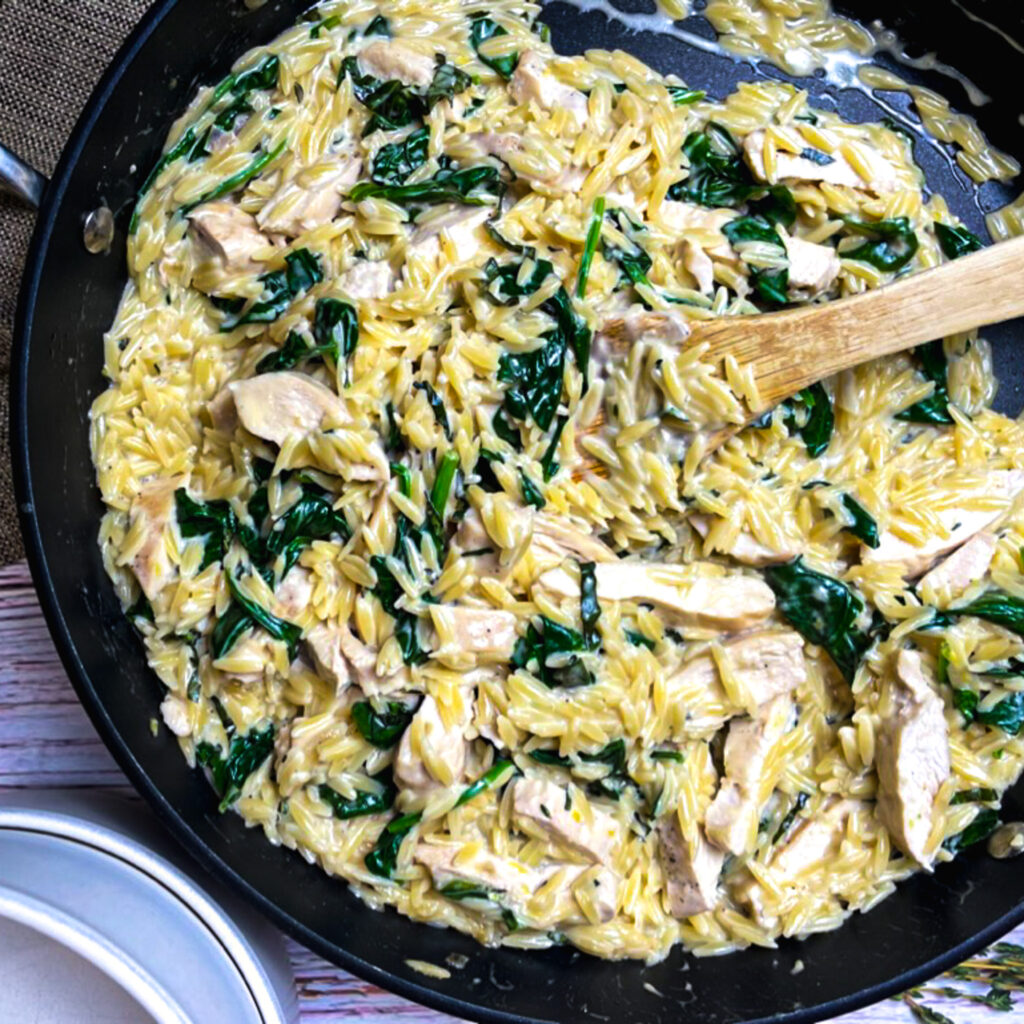 one pan lemon chicken orzo in a large fry pan with sliced chicken breast stirred in with a wooden serving spoon.
