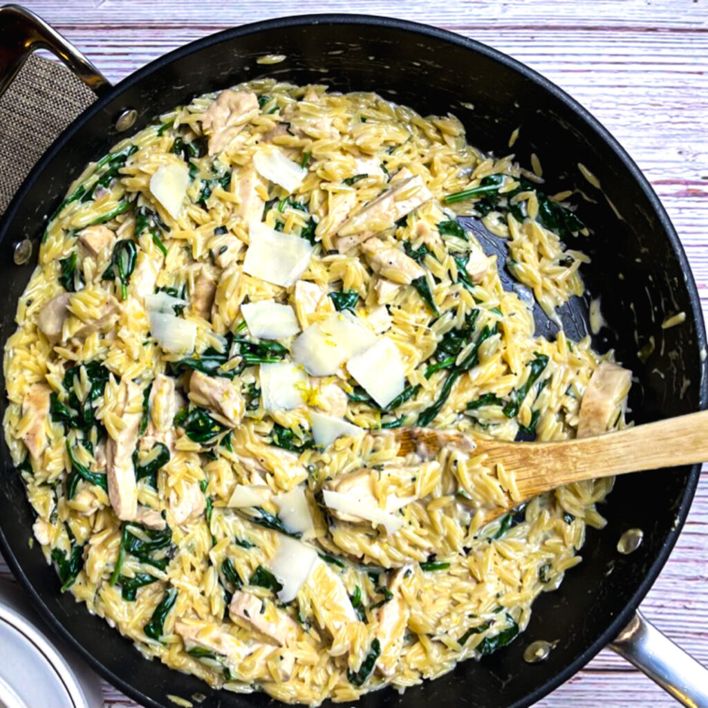 one pan lemon chicken orzo in a skillet with shaved parmesan and a bamboo serving spoon.