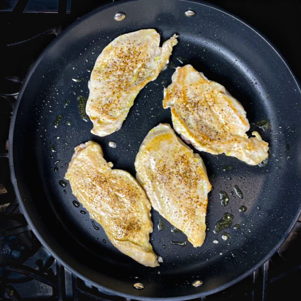 Four whole chicken breast frying in a large fry pan.