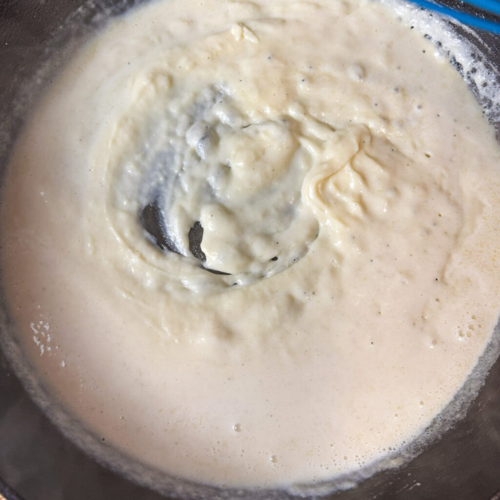 Homemade mac and cheese sauce thickening in a skillet. The smooth, creamy mixture is starting to come together as milk blends into the roux, creating a velvety base for the pasta.