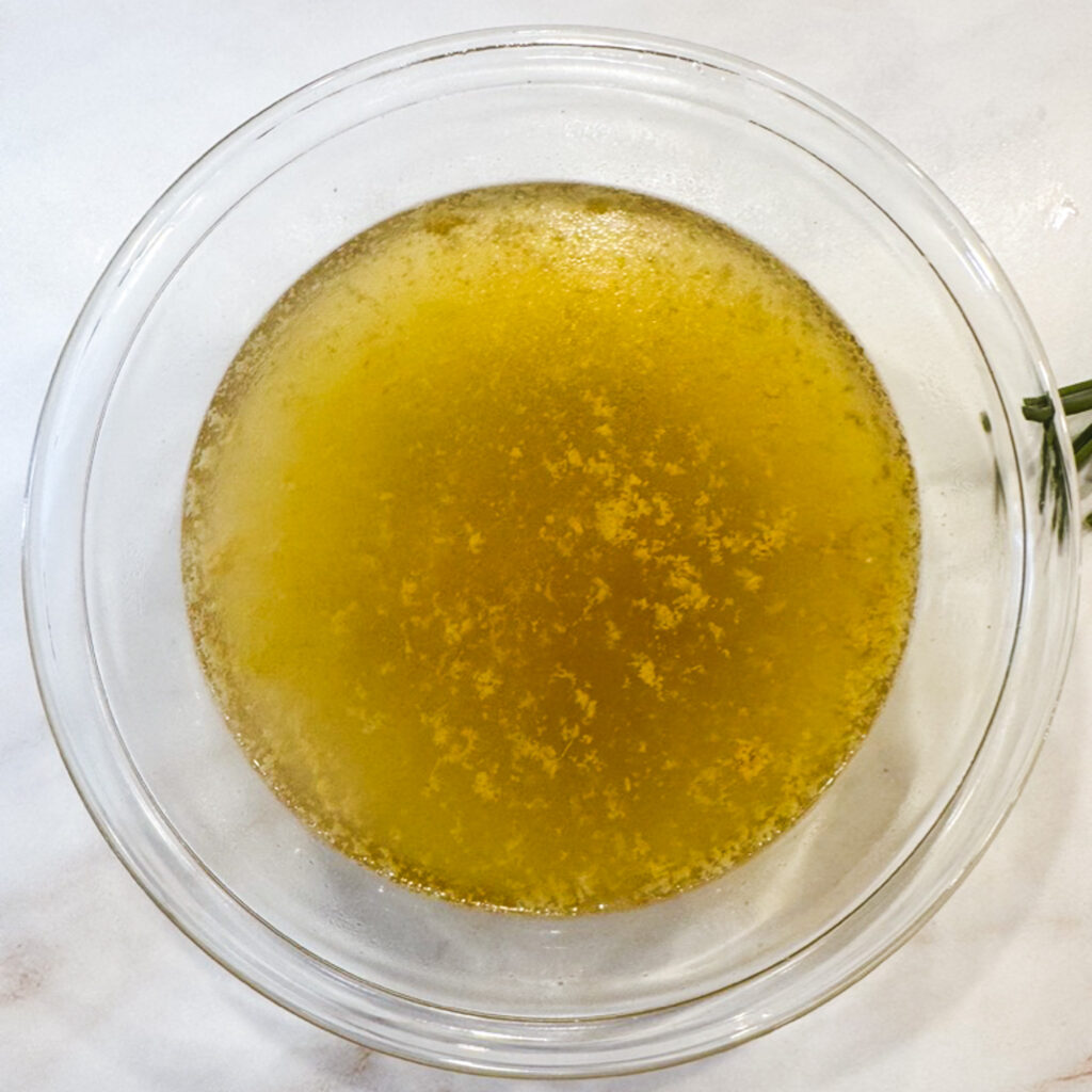 A glass bowl filled with rich, golden chicken broth after poaching — glistening and ready to be poured back into the pot.