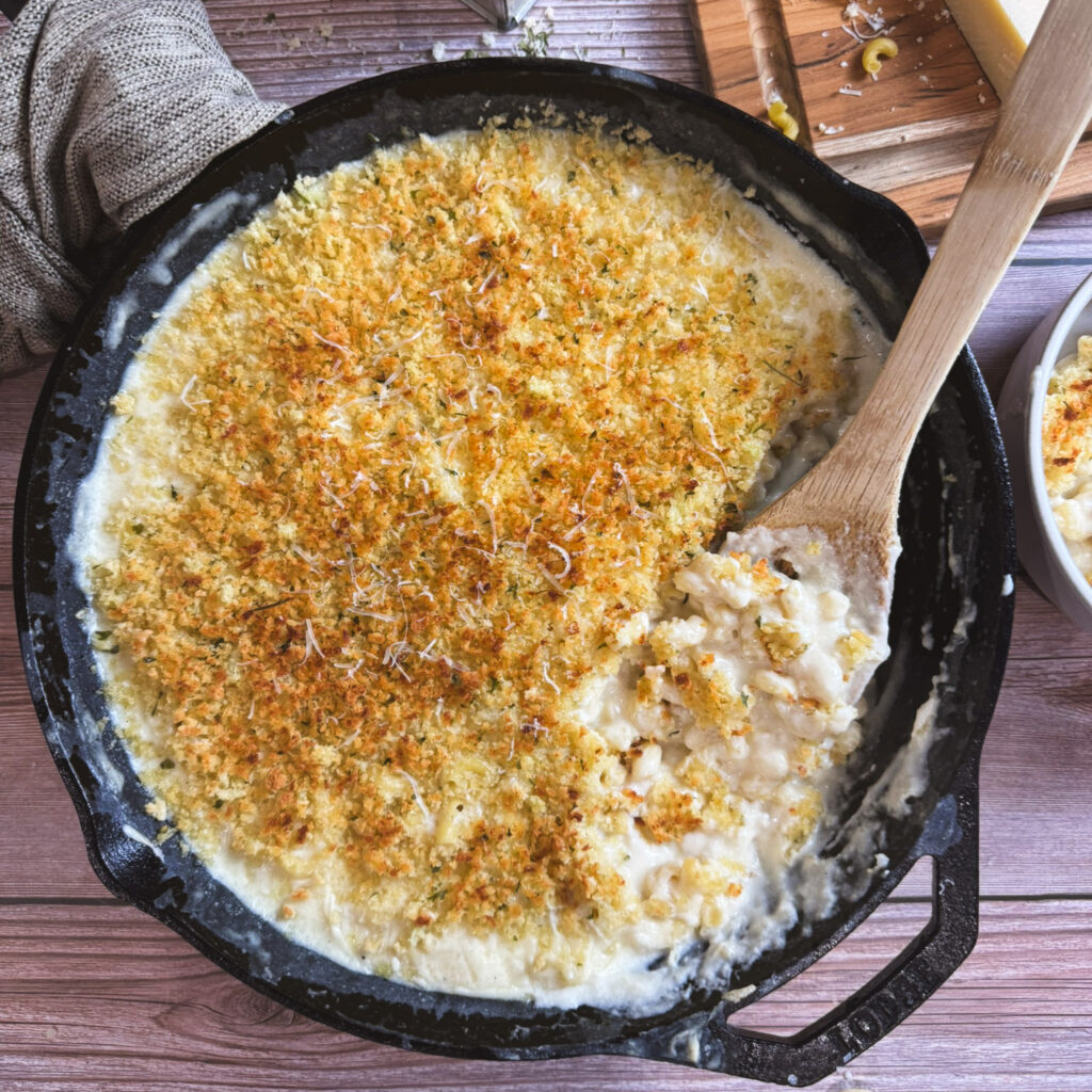 Overhead shot of a cast iron skillet filled with creamy baked mac and cheese. The golden breadcrumb topping is toasted and slightly uneven, with a wooden spoon lifting a serving to reveal the cheesy interior.