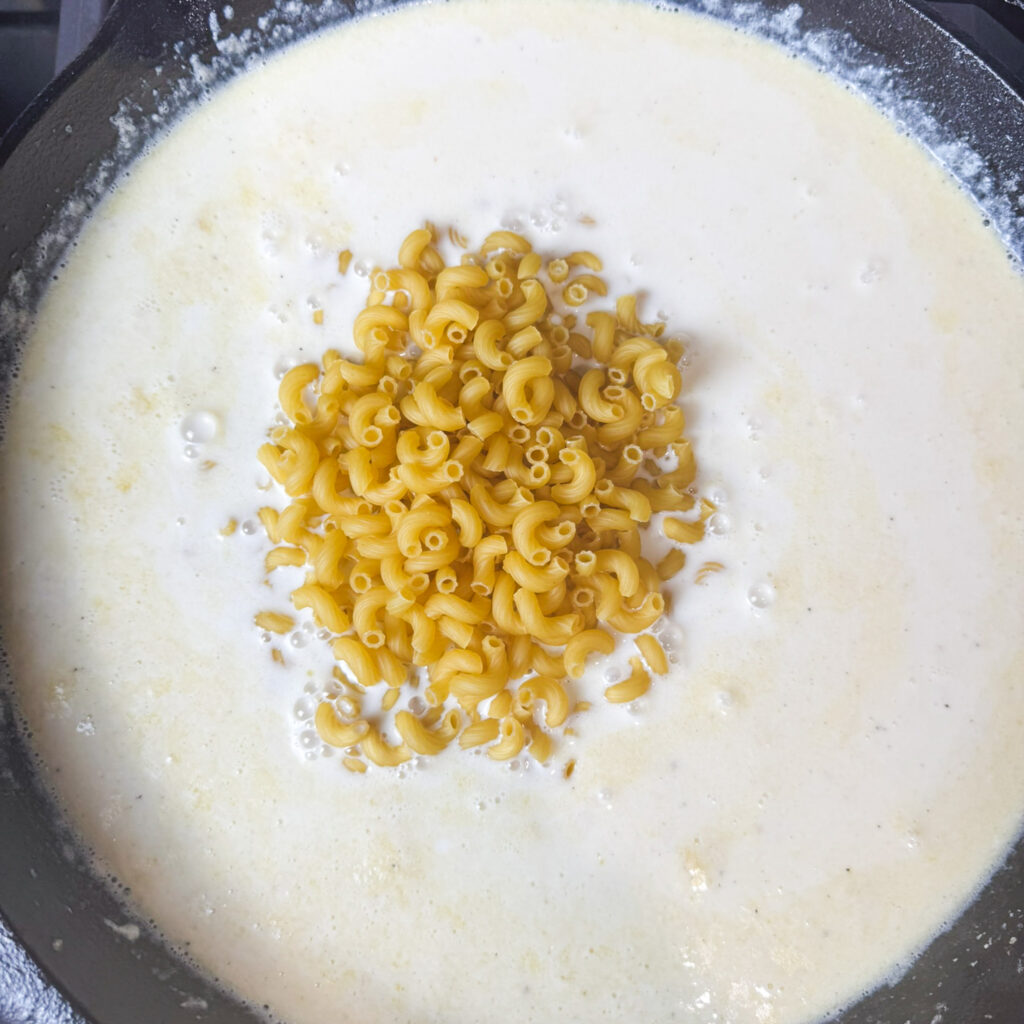 Uncooked elbow pasta added to a skillet of creamy milk and butter mixture — the first step in building this one-pot baked mac and cheese recipe.