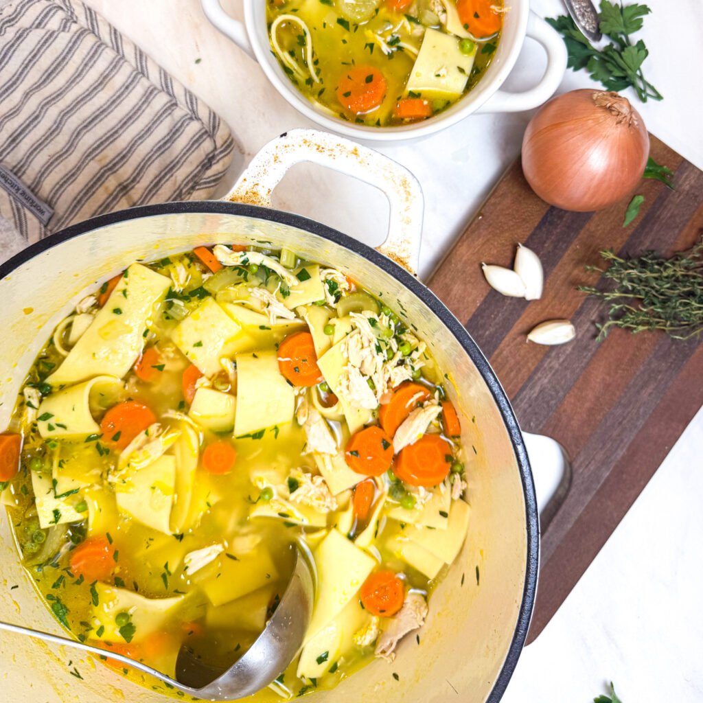 A cozy overhead view of chicken noodle soup in a white Dutch oven and a bowl beside it, filled with wide noodles, carrots, celery, peas, and shredded chicken in golden broth. Fresh herbs, a halved lemon, and a vintage spoon complete the warm, rustic scene.