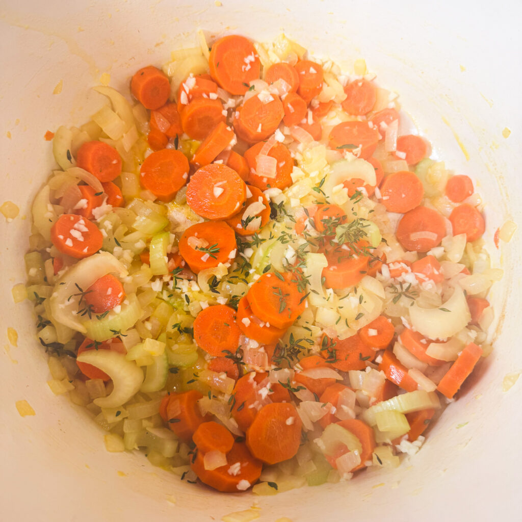 Chopped carrots, celery, and onion sautéing in a pot with minced garlic and fresh thyme, releasing fragrant steam as the base of the soup develops flavor.