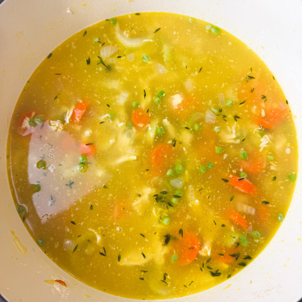 A pot of chicken noodle soup simmering with carrots, peas, and fresh herbs in golden broth — the flavors melding together beautifully.