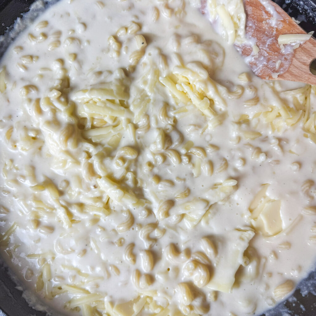 Shredded American cheese and cheddar melting into creamy pasta sauce in a cast iron skillet, creating the perfect gooey homemade mac and cheese.