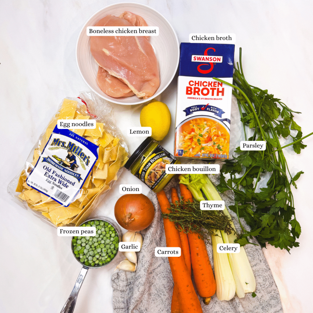 A flat lay of ingredients for homemade chicken noodle soup, including chicken breasts, egg noodles, carrots, celery, onion, garlic, parsley, thyme, lemon, chicken broth, chicken bouillon, and a measuring cup of frozen peas, all neatly arranged on a white surface.