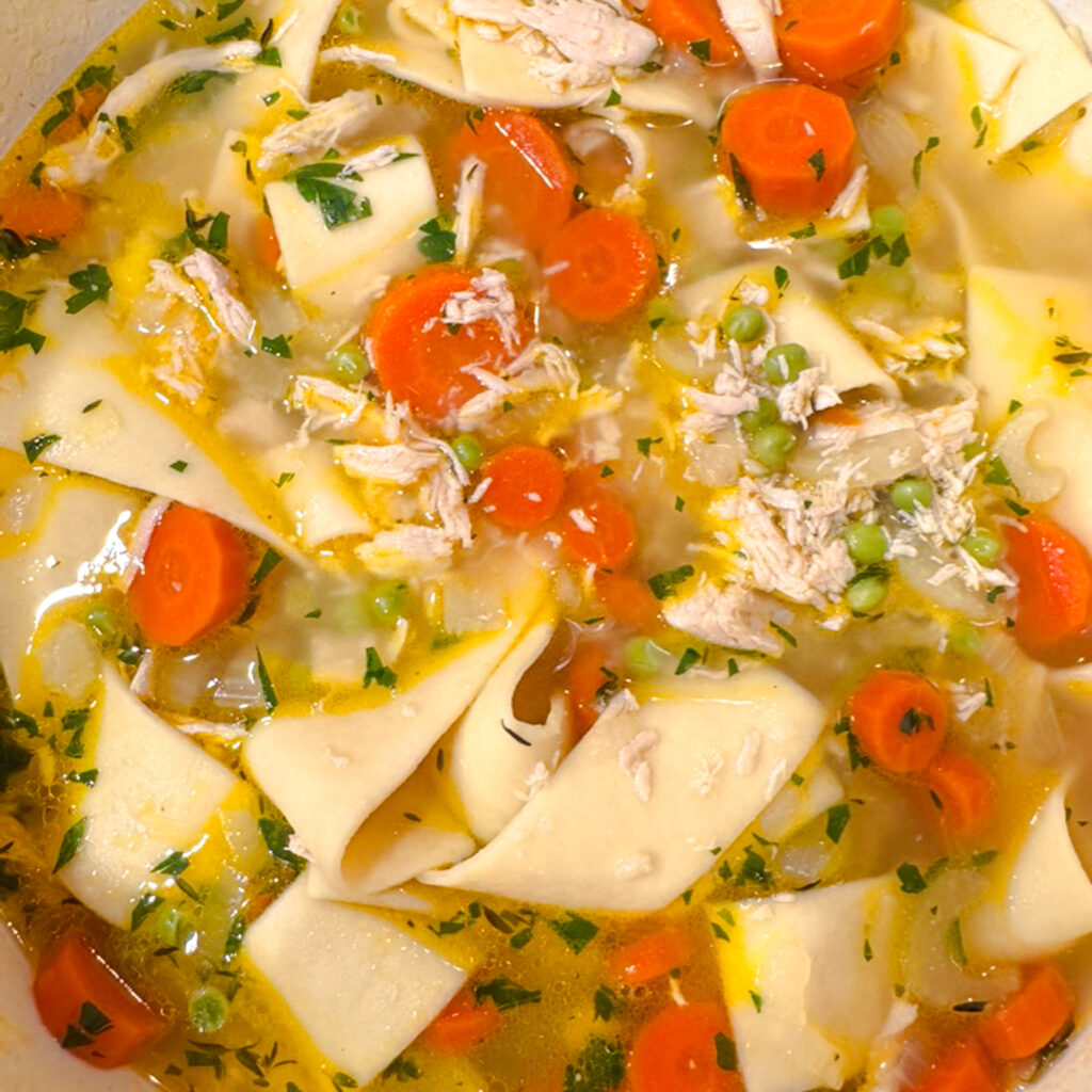 A close-up view of homemade chicken noodle soup showing tender wide egg noodles, shredded chicken, bright orange carrots, peas, and fresh herbs floating in a rich, golden broth.
